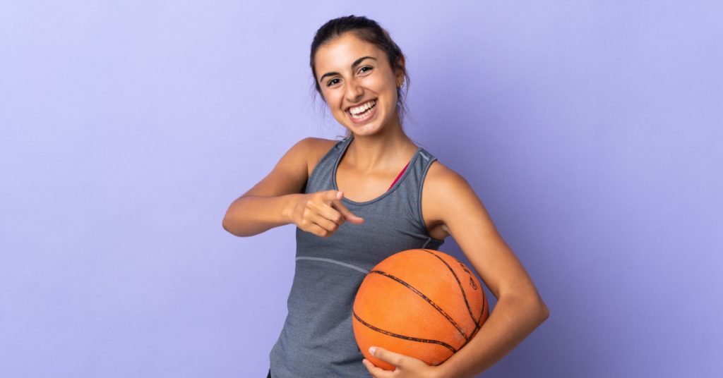 En la imagen aparecen una chica joven mirando hacia la cámara y señalando con un dedo hacia la misma, con amplia sonrisa, sosteniendo un balón de baloncesto con el otro brazo